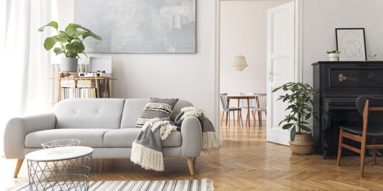 Modern scandianvian decor of living room with design sofa, elegant blanket, coffee table, plants and bookstand on the white wall. Design piano in the corner. Brown wooden parquet. View on dining room.